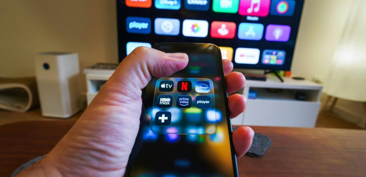 Close-up of a hand holding a phone displaying streaming apps in front of a TV with multiple app icons.
